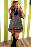 A woman takes a mirror selfie wearing a green, red, and blue plaid mini dress with black trim, paired with black knee-high boots, set against a green and white striped wall.