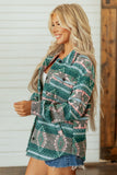 A woman wearing a long-sleeve button-up shirt with a green, pink, and gray geometric pattern, paired with denim shorts, standing in front of a white shiplap wall and wooden door.