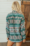 A woman viewed from behind wearing a long-sleeve patterned shirt with a green, gray, and pink geometric design, standing in front of a white siding and wooden door.