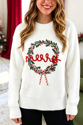 A woman wearing a white ribbed knit sweater with a green and red Christmas wreath and 'Merry' text graphic on the front, posed in a festive indoor setting.