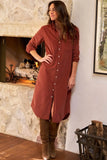 A woman wearing a rust-colored corduroy shirt dress with brown suede boots, standing in front of a stone fireplace.