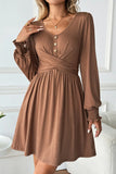 A woman wearing a brown long-sleeve V-neck dress with a buttoned henley-style chest, gathered waist, and smocked cuffs, shown in a bright indoor setting.