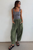 Woman wearing a gray tank top and green pants against a white wall.