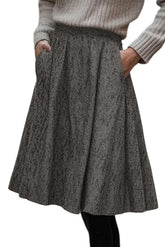 A gray and black herringbone patterned pleated skirt with side pockets, worn by a model in a ribbed beige sweater.