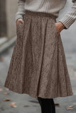A person wearing a brown and beige textured herringbone skirt with side pockets, paired with a light gray ribbed sweater and black leggings, standing outdoors on a paved street.