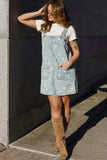 A woman wearing a light blue denim floral overalls dress over a white t-shirt and brown suede knee-high boots, posing outdoors against a dark gray concrete wall.