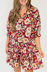 A woman wearing a short-sleeve collared dress with a red base and a large floral print in white, orange, and green, shown against a plain light gray background.