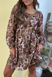 A woman wearing a brown floral long-sleeve mini dress with a square neckline and gathered waist, standing indoors next to a quilted chair.