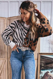 A woman models a cropped knit sweater featuring a split design: one half is black and white zebra print, and the other half is brown with black leopard spots. She is styled with blue jeans, a silver belt, and layered pearl necklaces, standing in front of a wooden screen.