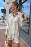 A woman wearing an oversized, light beige champagne-colored satin button-down shirt dress, standing outdoors on a brick sidewalk next to a storefront window.