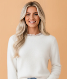 A smiling blonde woman wearing a soft white knit sweater with a delicate lace-trimmed crew neck, set against a solid peach-colored background.