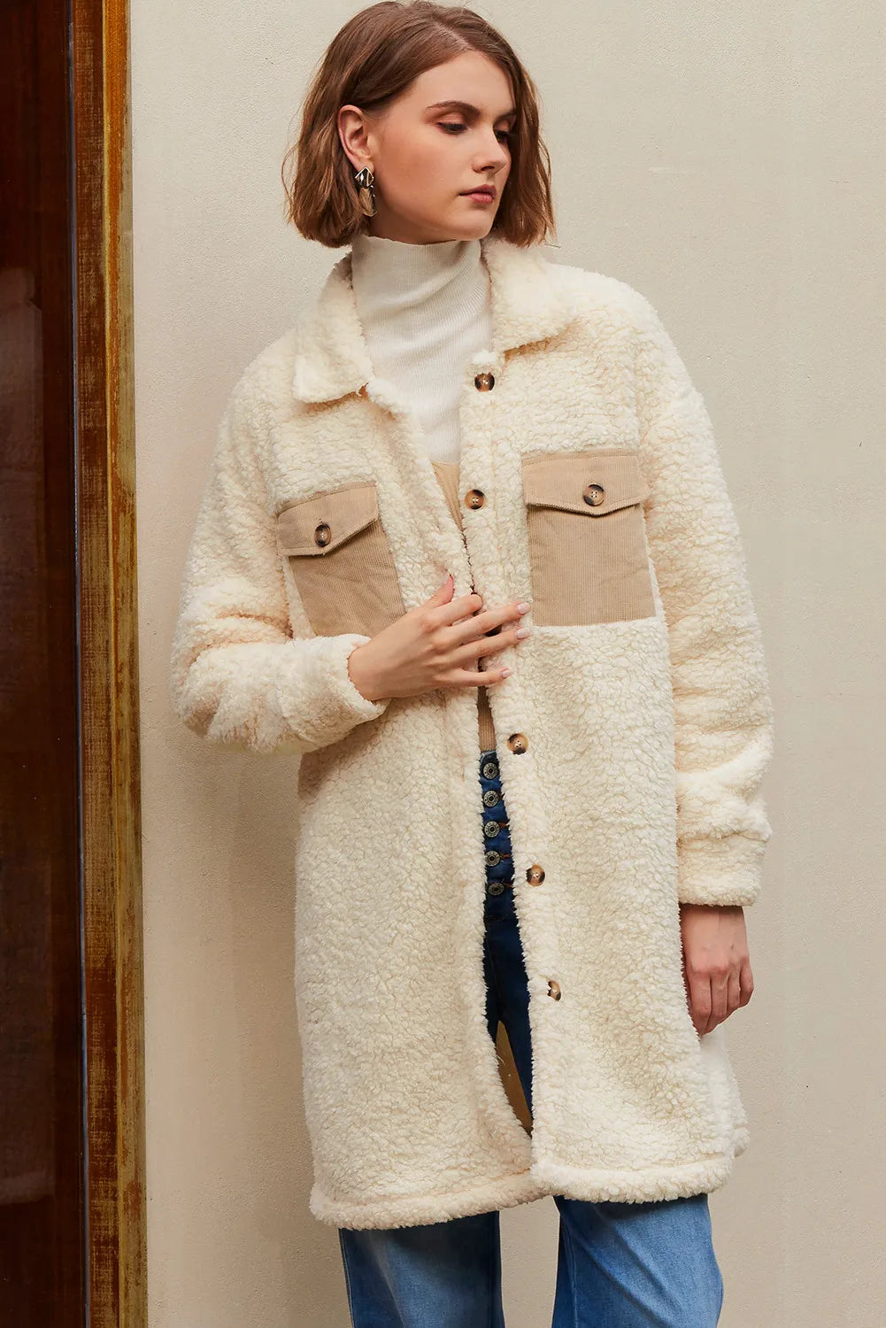 Woman in a cream sherpa coat, white turtleneck, blue jeans, against a beige wall.