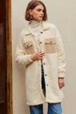 Woman in a cream sherpa coat, white turtleneck, blue jeans, against a beige wall.