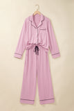 A light pink long-sleeve pajama set with dark piping trim, featuring a button-up front, chest pocket, and self-tie belt, hanging on a wooden hanger against a solid beige background.