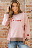 Woman wearing a pink sweatshirt with 'lover' and a heart design, standing against a brick wall.