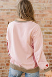 Person wearing a pink bomber jacket with a heart on the sleeve against a brick wall.