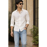Man wearing a white patterned shirt and light blue jeans standing outdoors.