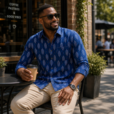 Man in a blue patterned shirt sitting outdoors holding a coffee cup.