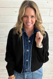 A smiling blonde woman wearing a black ribbed knit cardigan over a blue denim button-up shirt, paired with light blue jeans, standing against a white wood plank wall.
