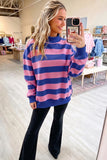 A woman takes a mirror selfie in a clothing boutique wearing a pink and blue horizontally striped mock neck sweatshirt and black flare pants.