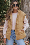 A woman wearing a tan quilted puffer vest over a light pink long-sleeve top and blue jeans, standing outdoors in front of a tree trunk.