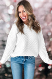 A smiling woman wearing a white textured knit sweater and blue jeans, standing against a soft pink bokeh background.