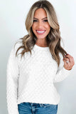 A smiling woman with long highlighted brown hair wearing a white textured knit sweater and blue jeans, standing against a light gray studio background.