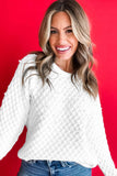 A smiling woman with blonde-highlighted hair wearing a white textured knit sweater with pearl-like embellishments and blue jeans, posed against a solid bright red background.