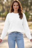 A woman wearing a white textured knit long-sleeve sweater and light blue jeans, standing outdoors with a blurred natural background.