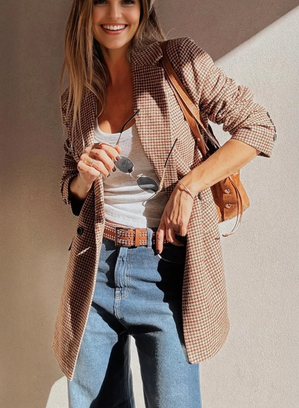 Woman wearing a plaid jacket, white top, and blue jeans with a brown bag against a neutral background