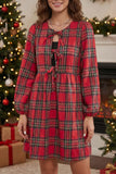 A woman wearing a red plaid long-sleeve dress with a tie-front neckline and gathered waist, standing in a holiday-themed room with Christmas trees and wrapped gifts in the background.
