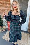 A woman wearing a long-sleeve plaid midi dress in dark green and navy, standing on a porch in front of a brick wall and white door.