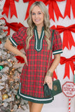 A woman modeling a red and green plaid holiday tunic dress with white trim, standing in front of a white wall decorated with red bows and a Christmas tree.