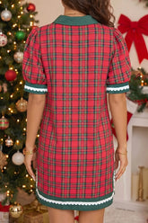 A woman wearing a red and green plaid short-sleeve dress with a green collar and white scalloped trim, photographed from the back in a holiday-themed room with a Christmas tree and fireplace in the background.