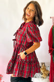 A woman modeling a red and black plaid short-sleeve tunic dress with tie details on the chest and a gathered waist, standing against a light pink wall.