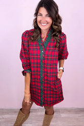 A woman wearing a red and green plaid button-down shirt dress with three-quarter sleeves and brown boots, standing against a light pink wall.