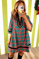 A woman takes a mirror selfie wearing a festive plaid tunic dress with red, green, and white stripes and black trim, set against a lime green and white striped wall.