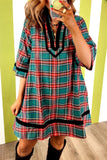 A woman takes a mirror selfie wearing a short-sleeve plaid tunic dress with a teal, red, and white pattern and black trim. She is wearing black boots and standing in front of a green and white striped wall.