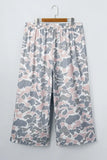 A pair of wide-leg cropped pants with an elastic waistband, featuring a pink, gray, and white camouflage pattern, hanging on a wooden hanger against a light gray wall.