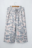 Wide-leg cropped pants with a gray, white, and dusty pink camouflage pattern, hanging on a wooden hanger against a light gray wall.