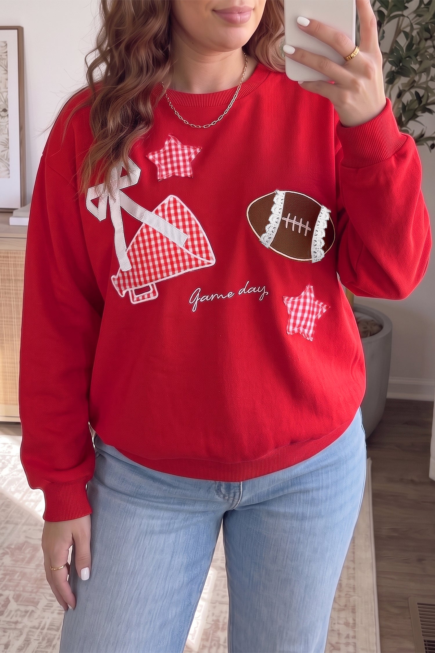 Woman wearing a red football graphic sweatshirt with light blue jeans, taking a mirror selfie.