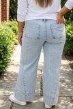 A person viewed from behind wearing light-wash wide-leg denim jeans, a white long-sleeve top, and white sneakers, standing on a stone path outdoors with greenery on both sides.