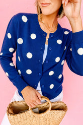 A woman wearing a navy blue cardigan with large white polka dots, holding a woven straw handbag against a solid pink background.