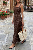 Woman in a brown polka dot dress holding a straw bag on a stone street with flowers and tables in the background.