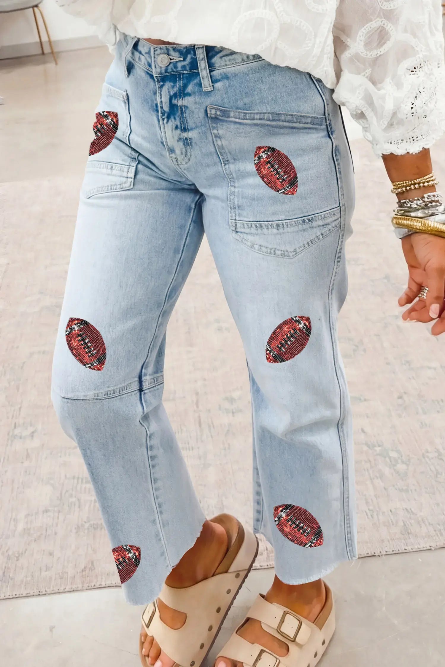 Light wash blue denim jeans with embroidered football patches on the thighs and lower legs, shown on a model wearing a white lace-trim top and beige slide sandals.