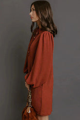 A woman in side profile wearing a rust-orange long-sleeve blouse with puffed shoulders, holding a brown handbag with a gold chain strap, set against a solid dark gray background.