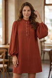 A woman wearing a rust-colored long-sleeve dress with a ruffled high neck and button placket, standing in a softly lit indoor setting with wooden furniture and windows in the background.