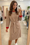 A woman takes a mirror selfie in a clothing boutique wearing a brown and cream striped long-sleeve dress with red cuffs and a red crew neck, paired with white sneakers.