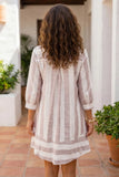 A woman viewed from behind wearing a light pink and white vertically striped linen tunic dress with three-quarter sleeves, set against a warm, sunlit outdoor patio background.