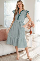 A woman modeling a green and white vertically striped midi dress with ruffled cap sleeves, a split neckline, and a tiered skirt, standing in a bright, modern living room.